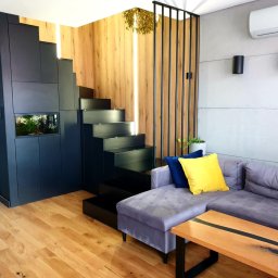 BARTOSZ MROCZEK BARTMEB - Nowoczesne wnętrze z czarnymi schodami prowadzącymi na antresolę, wkomponowanymi w zabudowę meblową z akwarium, drewnianą ścianą z podświetleniem i szarą sofą z kolorowymi poduszkami.