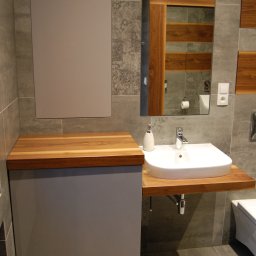 BARTOSZ MROCZEK BARTMEB - Szara szafka łazienkowa z drewnianym blatem, na którym osadzona jest biała umywalka. Nad szafką wisi lustro z drewnianą ramą, a obok widać fragment toalety. Na ścianach szare płytki.