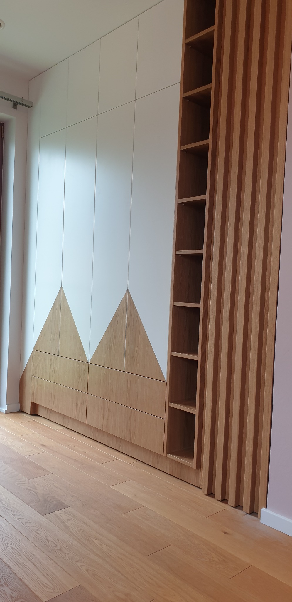 Biało-drewniana szafa wnękowa z geometrycznym wzorem trójkątów i regałem z listew, widok z dołu na parkiet.