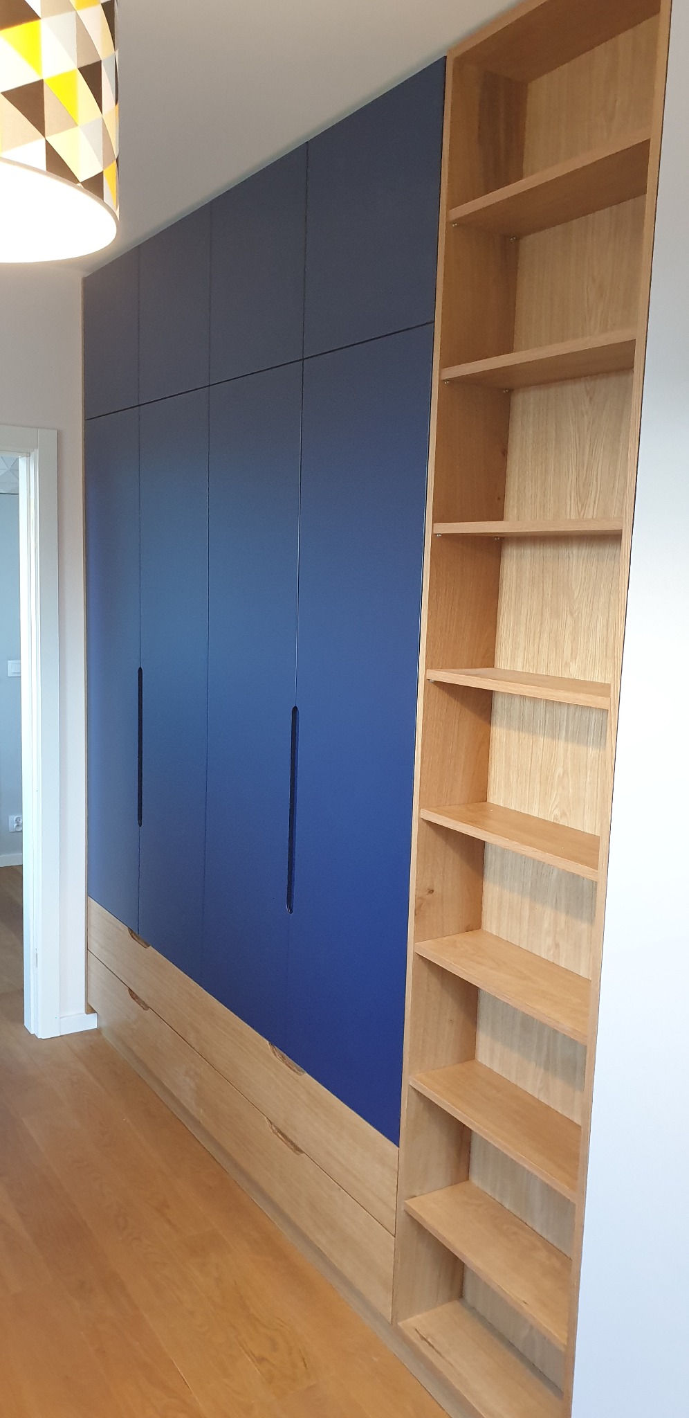 Niebieska zabudowa meblowa z szafkami i drewnianymi półkami, zintegrowana z podłogą, oświetlona lampą z geometrycznym wzorem.