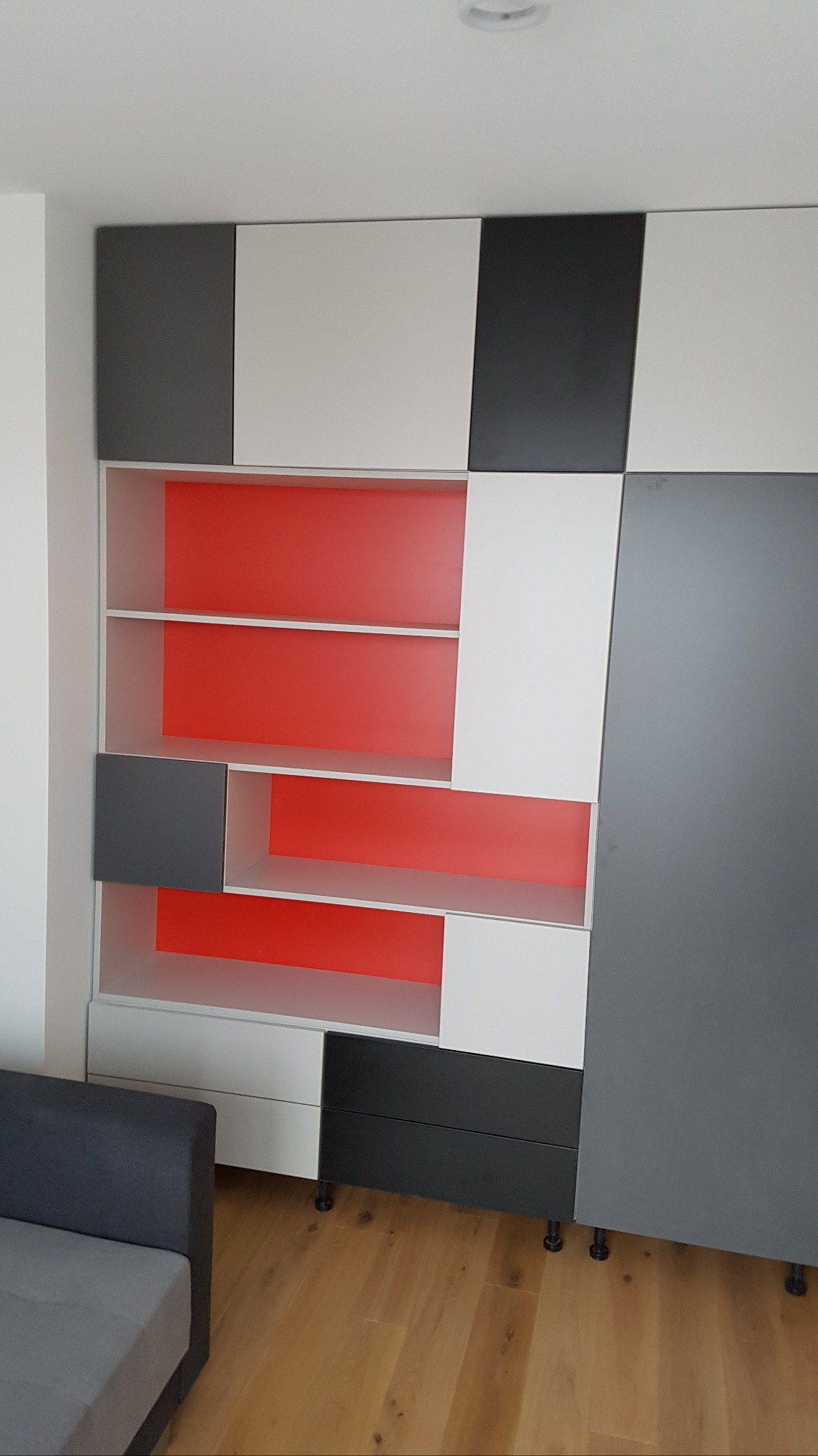 Nowoczesna szafa wnękowa z białymi, szarymi i czarnymi frontami, z czerwonymi wstawkami w otwartych półkach, widoczne małe nóżki szafki.
