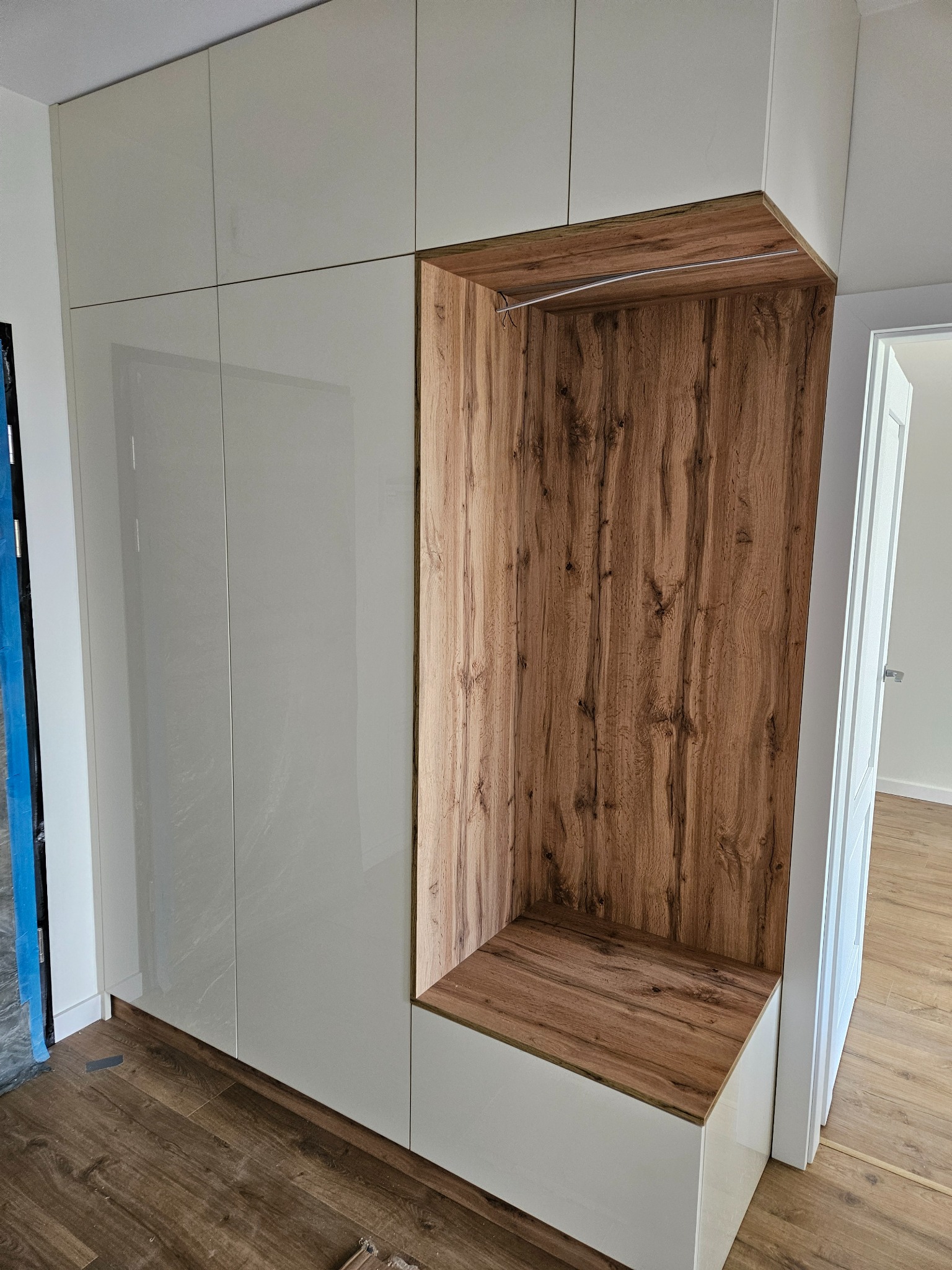 Biała szafa wnękowa z drewnianą wnęką na buty i wieszak, widok z boku na parkiet i uchylone drzwi do pokoju.