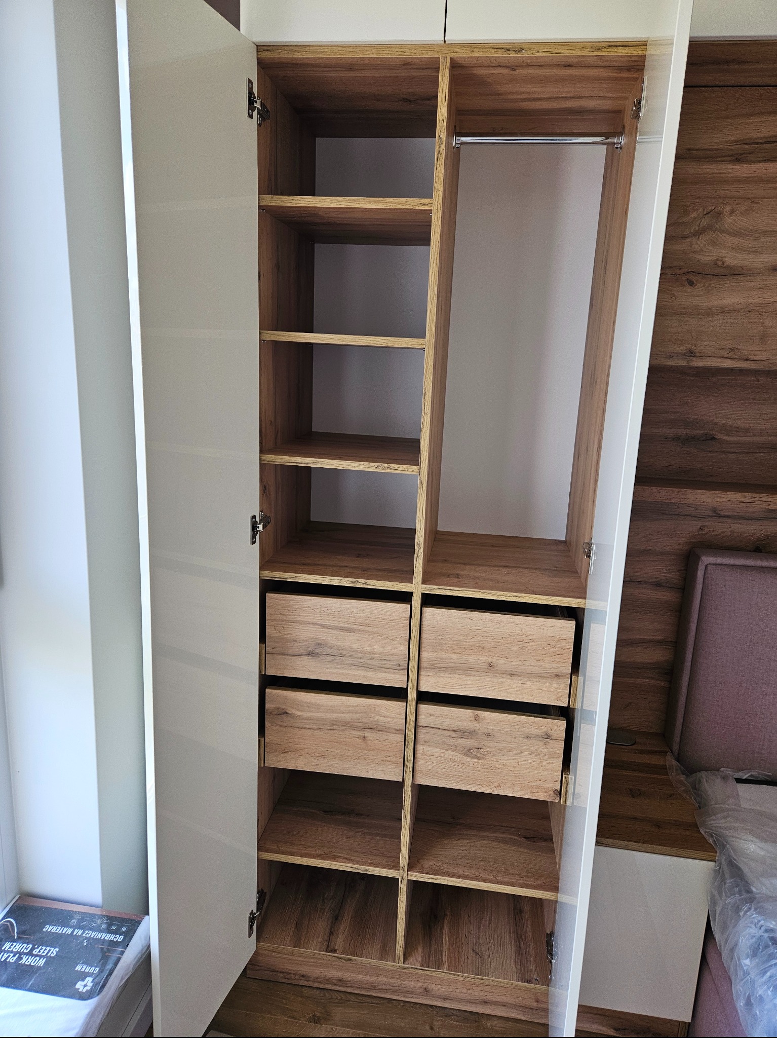 Otwarta szafa z białymi frontami i drewnianymi elementami wewnętrznymi: półkami, szufladami i drążkiem na ubrania. Widoczny fragment sypialni z łóżkiem i szafką nocną.