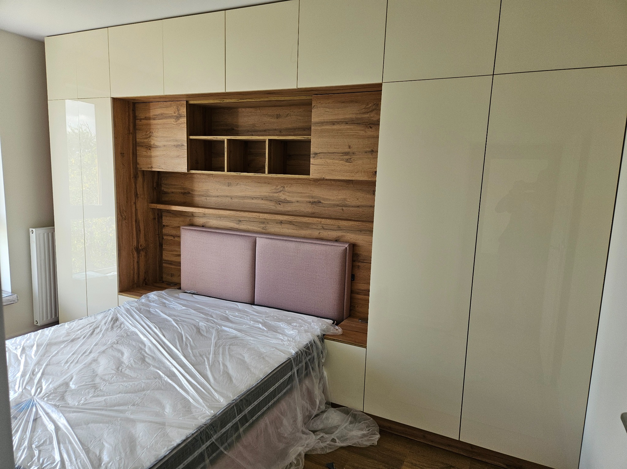 Nowoczesna sypialnia z zabudową meblową: beżowe szafy z połyskiem integrują się z drewnianą wnęką nad łóżkiem z różowym zagłówkiem; materac zabezpieczony folią ochronną.