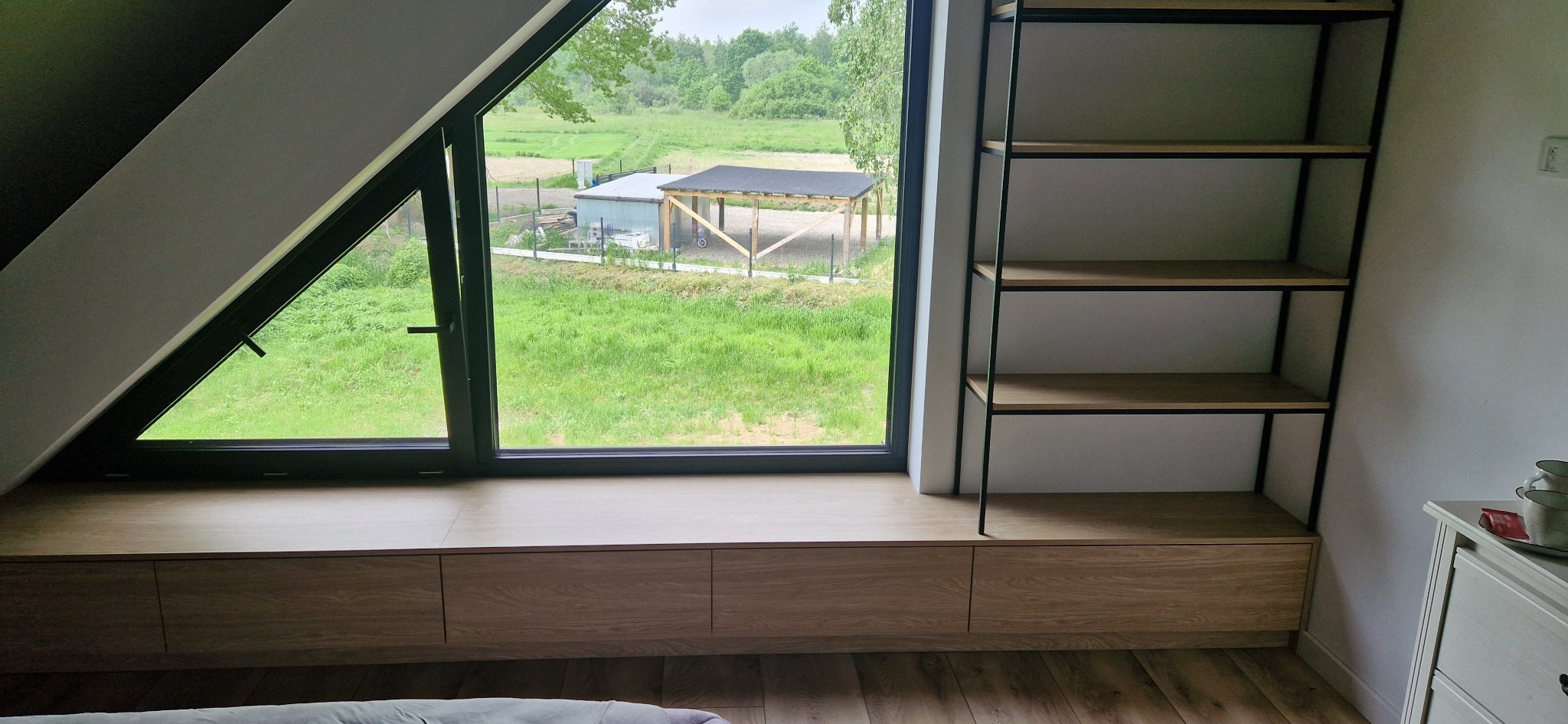 Wnęka okienna z zabudową z drewna dębowego i czarne metalowe półki w pokoju na poddaszu z widokiem na zielony krajobraz.