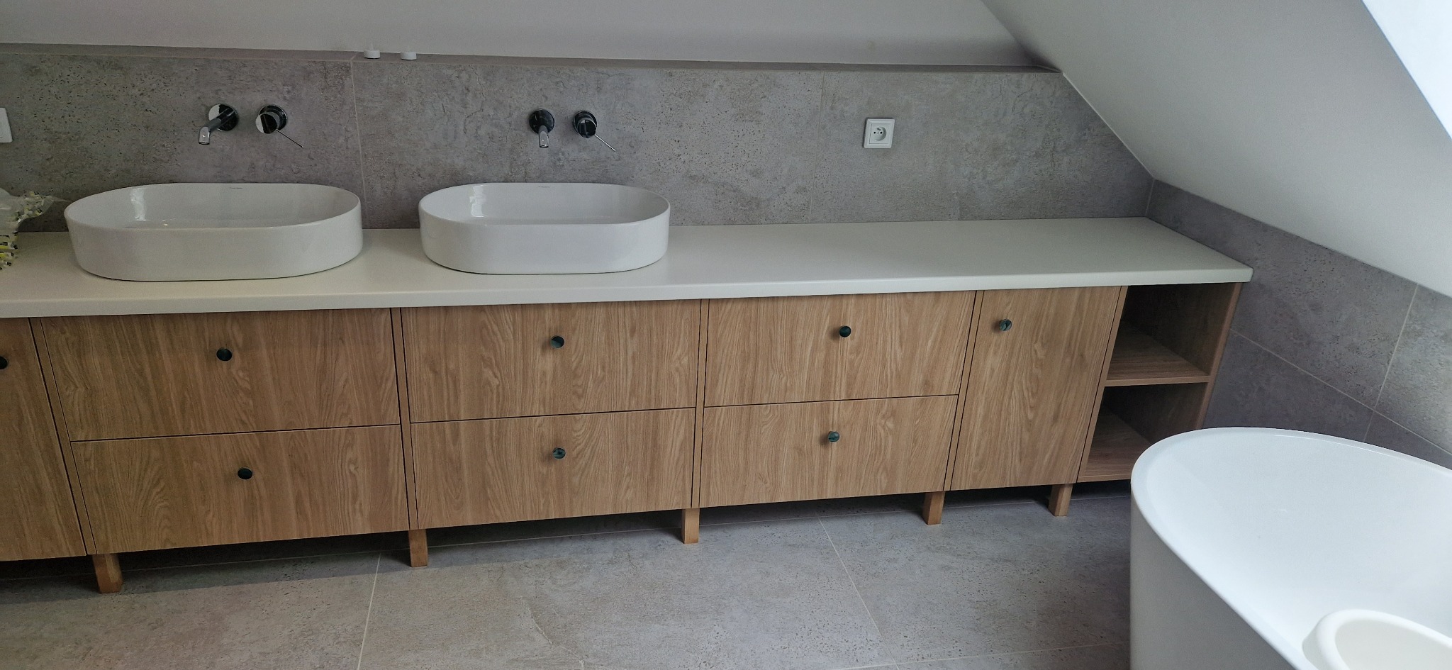 Łazienka w skandynawskim stylu z drewnianą szafką pod umywalki nablatowe i szarą płytką imitującą beton, oświetlona naturalnym światłem z okna dachowego.