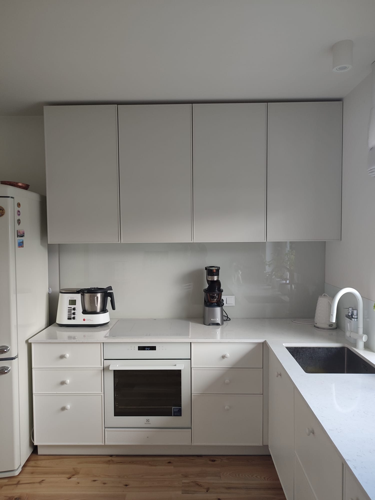 Jasna kuchnia z białymi, gładkimi frontami szafek wiszących i dolnych, białym blatem z szarą żyłką, sprzęt AGD na blacie: robot kuchenny, wyciskarka wolnoobrotowa i czajnik elektryczny.