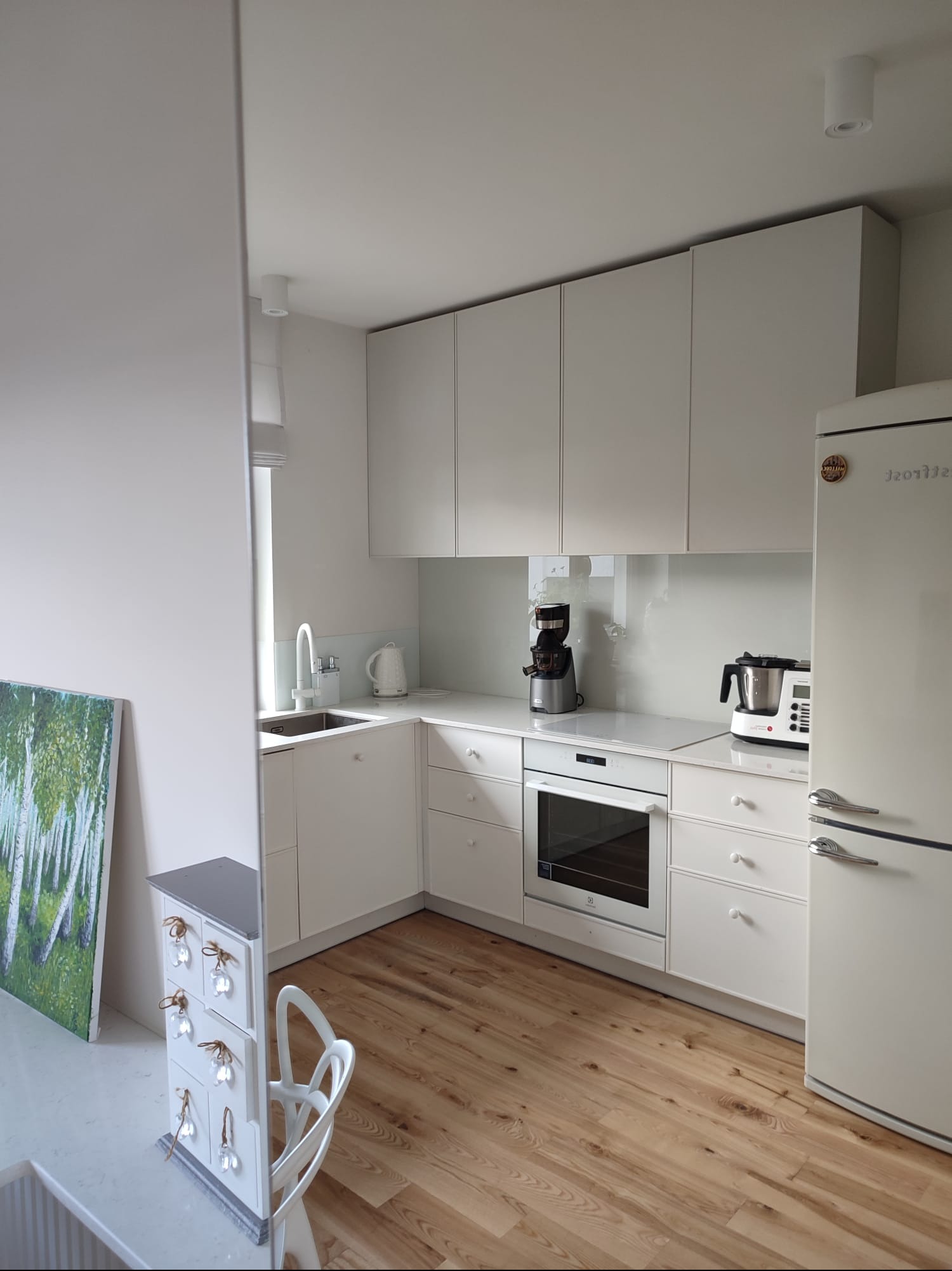 Jasna kuchnia w stylu skandynawskim z drewnianą podłogą, białymi szafkami bez uchwytów, lodówką retro i elementami dekoracyjnymi, takimi jak obraz z motywem leśnym i mała komoda z kryształowymi...