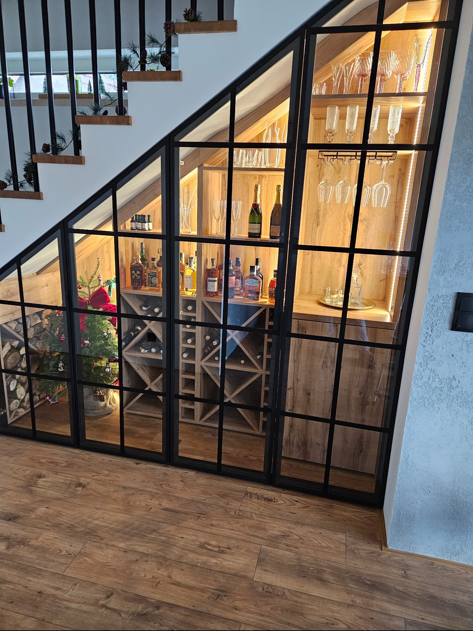 Zabudowa wnęki pod schodami z czarną, metalową ramą i szklanymi drzwiczkami, tworząca barko-spiżarnię z oświetleniem LED, butelkami alkoholu, kieliszkami i małą choinką.