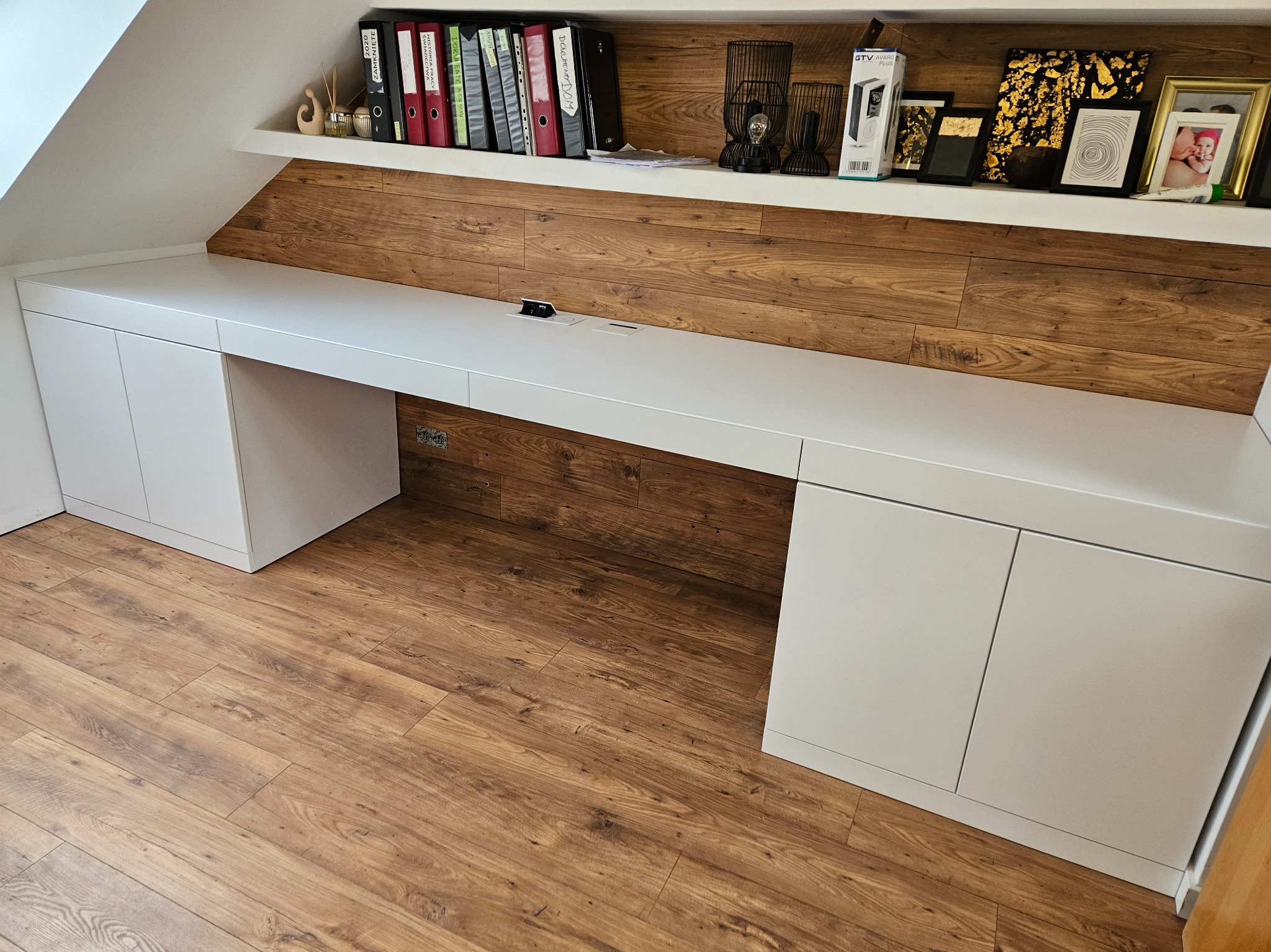 Białe biurko na wymiar wkomponowane w skos poddasza, z szafkami po bokach i drewnianą boazerią na ścianie, uzupełnione półką z dekoracjami i segregatorami.