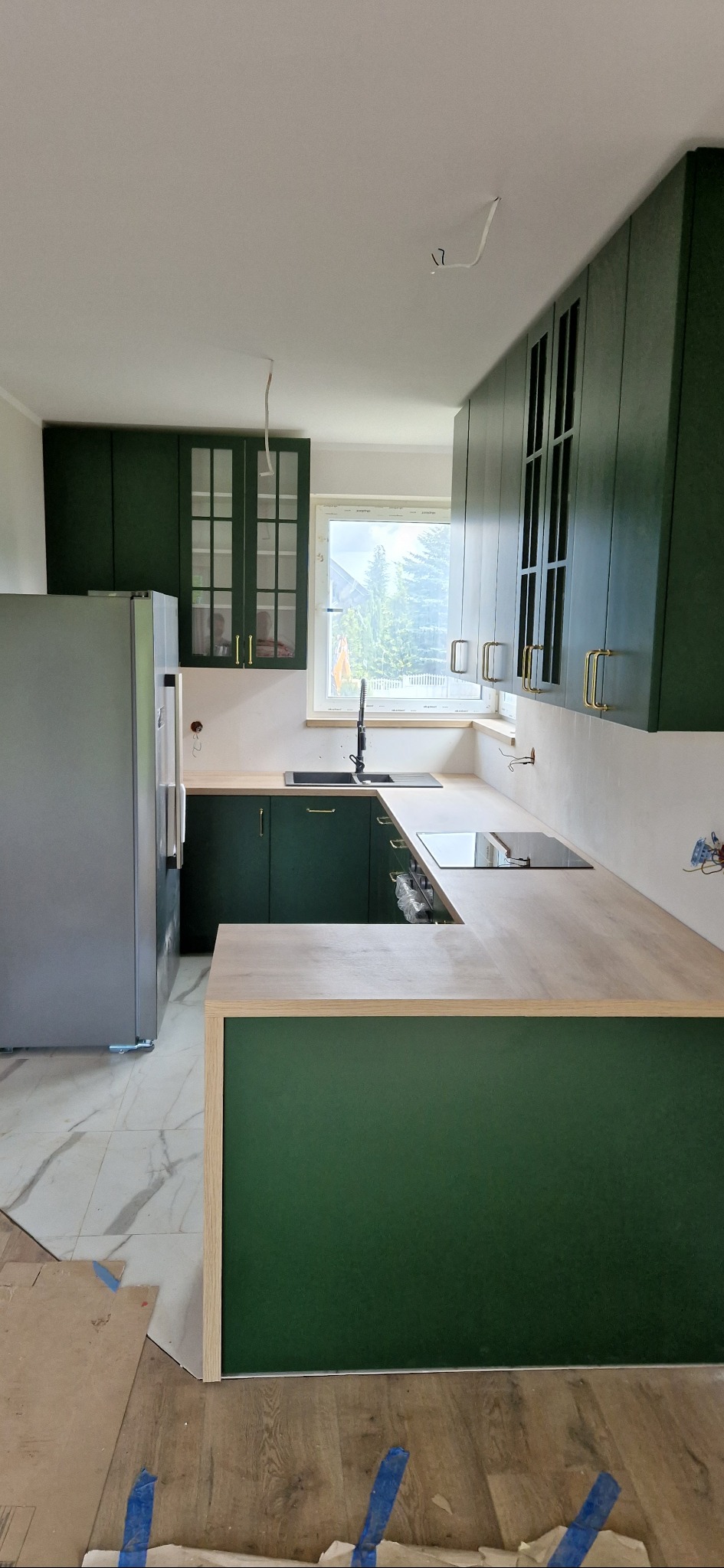 Nowoczesna kuchnia w odcieniach zieleni z drewnianym blatem, widoczne niedokończone prace wykończeniowe na podłodze i ścianach, okno z widokiem na zieleń za nim.