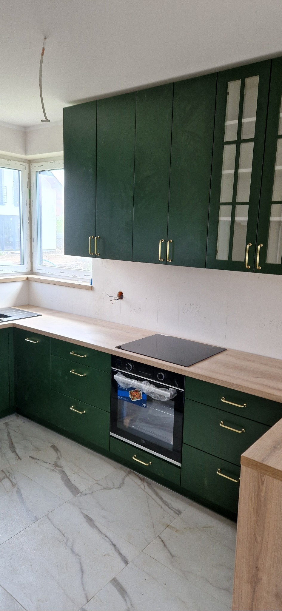 Nowoczesna kuchnia z ciemnozielonymi szafkami i złotymi uchwytami, jasny blat, płyta indukcyjna i piekarnik w folii, marmurowa podłoga, widoczne niedokończone prace elektryczne