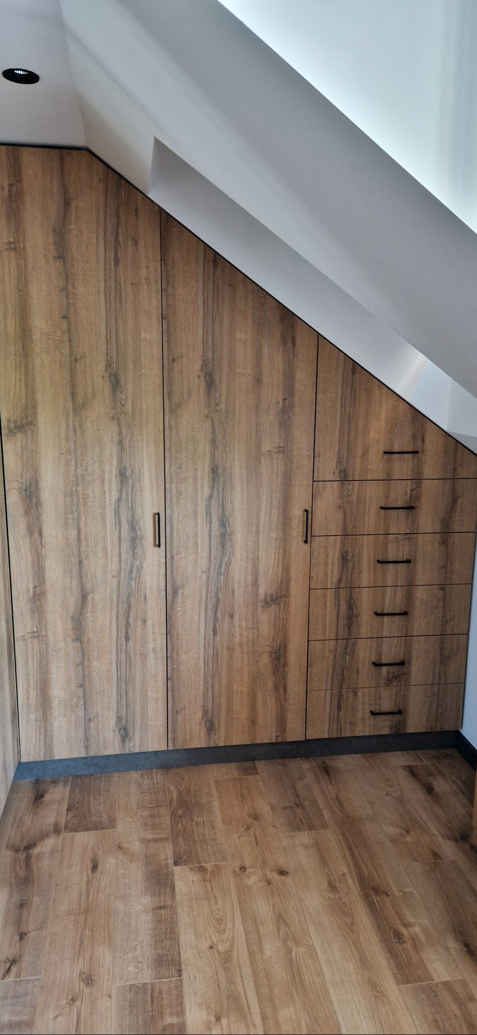 Zabudowa wnęki na poddaszu z drewnopodobnymi frontami, czarnymi uchwytami i czterema szufladami, dopasowana do skosu dachu.