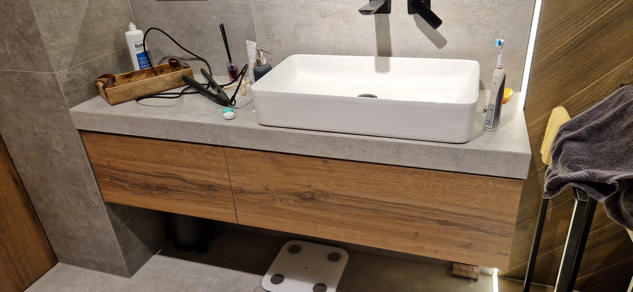 Nowoczesna łazienka z drewnianą szafką pod umywalką, na której stoją przybory toaletowe, w tle widoczna waga łazienkowa.