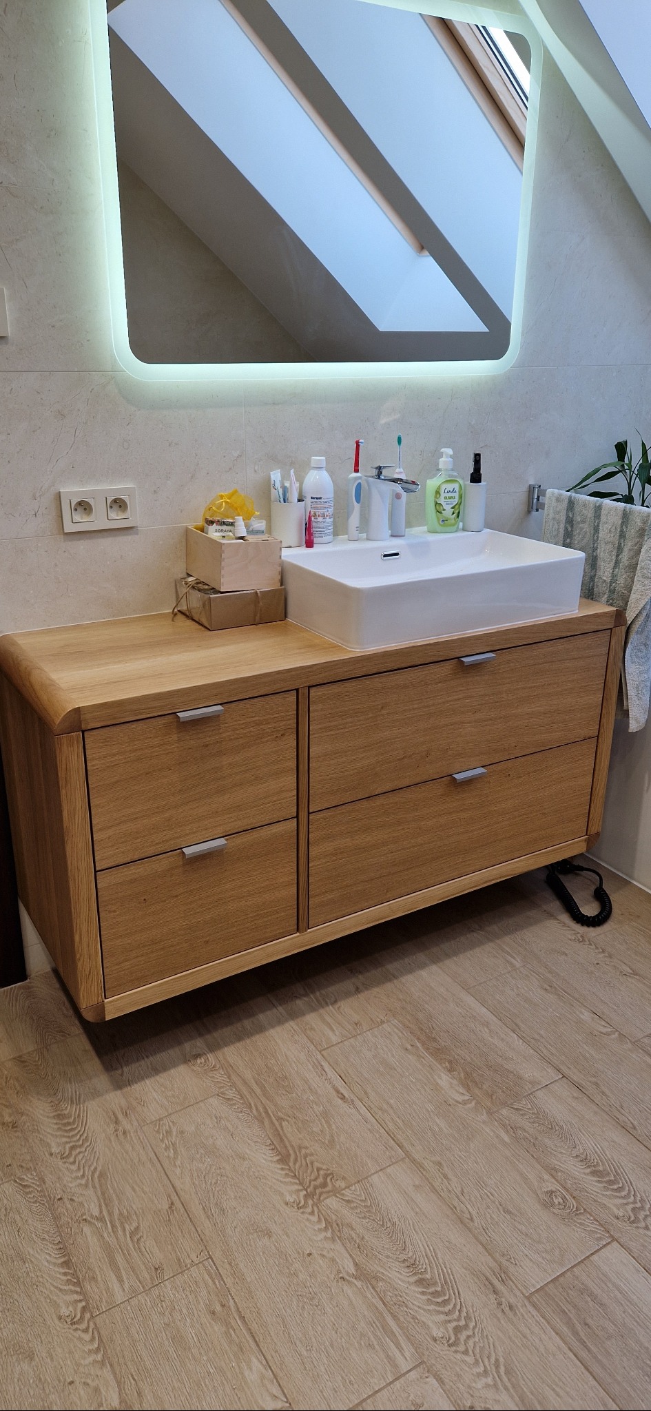 Nowoczesna łazienka z drewnianą szafką pod umywalką, białą umywalką nablatową i podświetlanym lustrem, widok na okno dachowe.