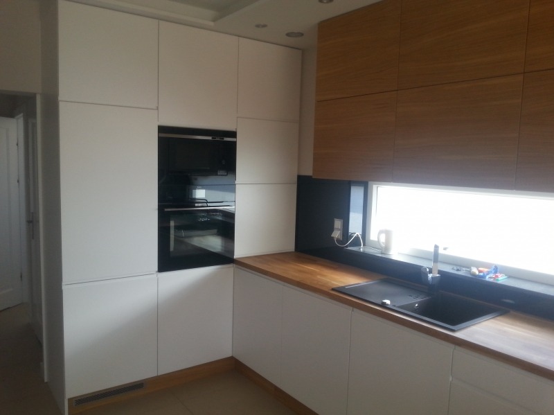 Nowoczesna kuchnia z białymi szafkami, drewnianym blatem i czarnym zlewem pod oknem, widoczny piekarnik w zabudowie.