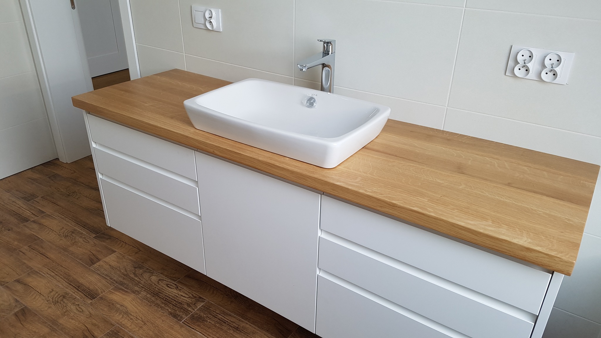 Nowoczesna łazienka z białą szafką pod umywalkę z drewnianym blatem, prostokątna umywalka nablatowa i chromowana bateria, białe płytki na ścianie i drewnopodobna podłoga.