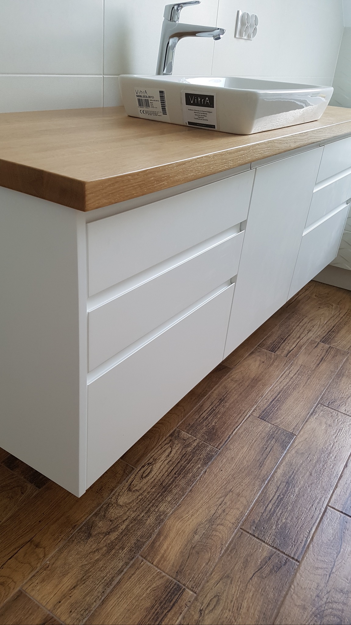 Biała szafka łazienkowa z drewnianym blatem, umywalką nablatową marki Vitra i chromowaną baterią, na tle białych płytek i drewnopodobnej podłogi.