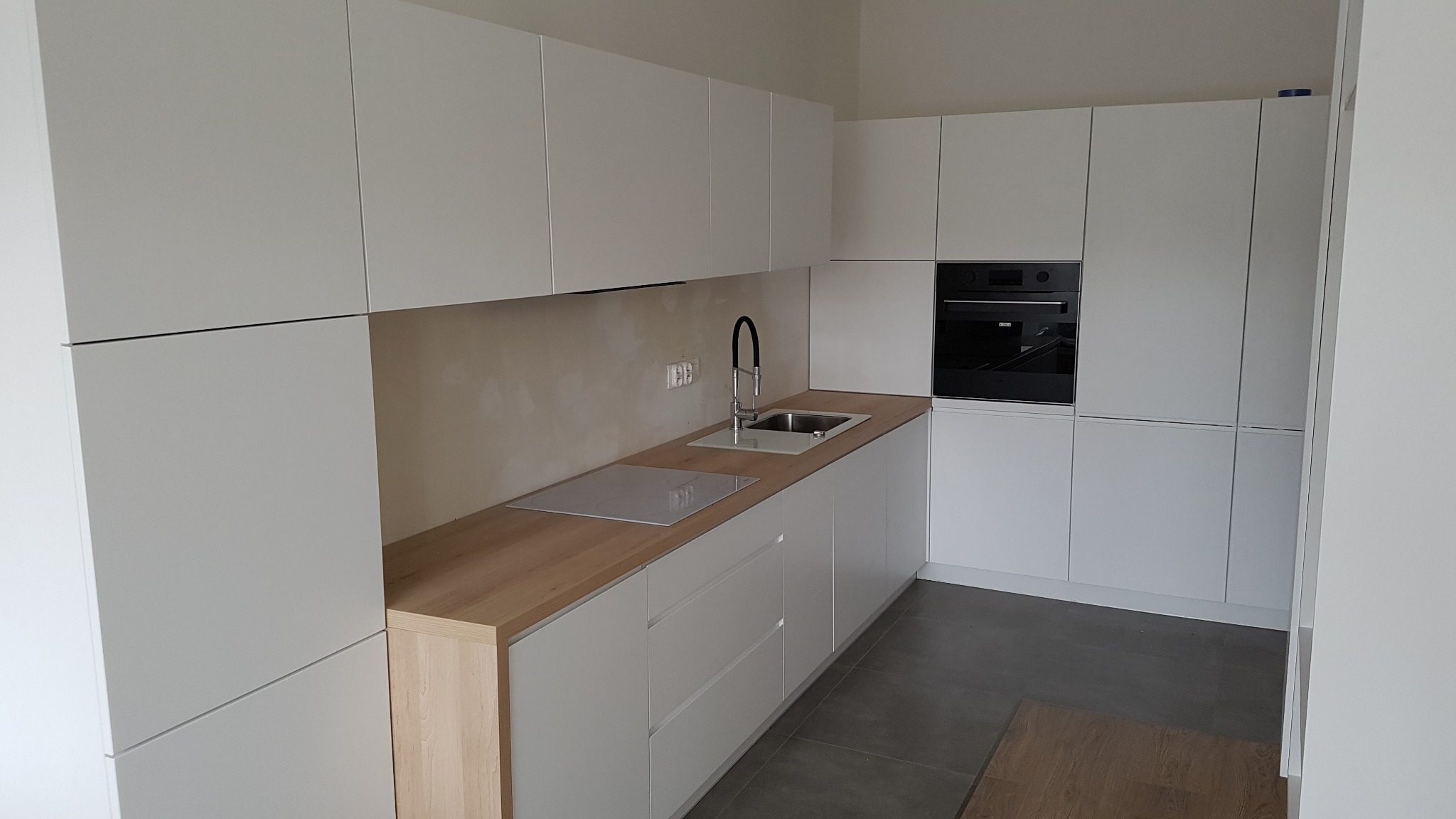 Nowoczesna, minimalistyczna kuchnia z białymi szafkami i drewnianym blatem, widoczny zlew z czarną baterią i płyta indukcyjna, szare płytki na podłodze.
