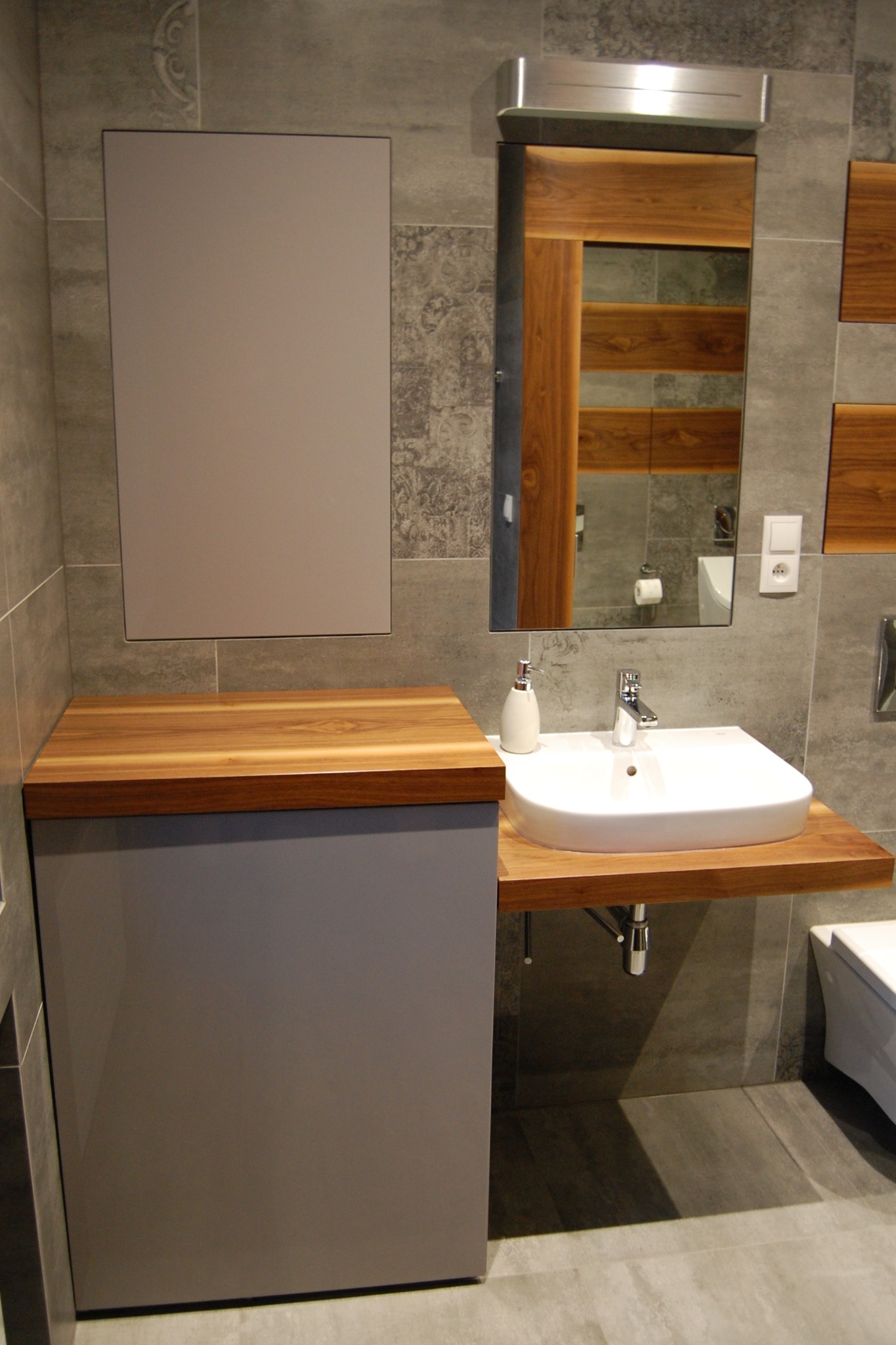 Szara szafka łazienkowa z drewnianym blatem, na którym osadzona jest biała umywalka. Nad szafką wisi lustro z drewnianą ramą, a obok widać fragment toalety. Na ścianach szare płytki.