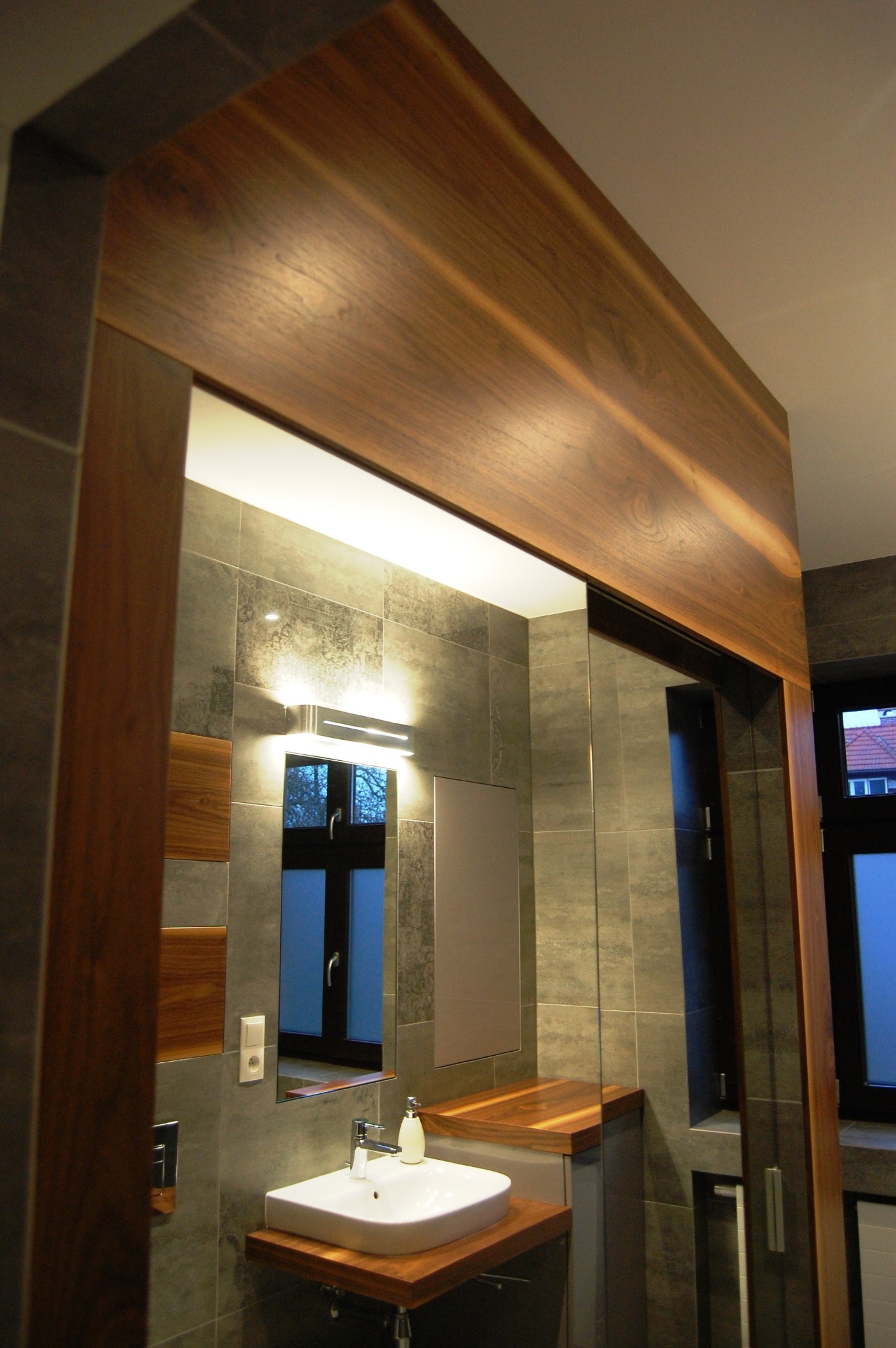 Nowoczesna łazienka z drewnianymi akcentami: obudowa lustra i blaty z ciemnego drewna, szare płytki, biała umywalka i minimalistyczne oświetlenie.