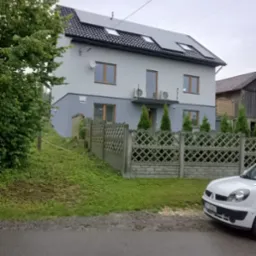 Dwukondygnacyjny dom z szarą elewacją, panelami słonecznymi na dachu i betonowym ogrodzeniem, widok z ulicy w pochmurny dzień.