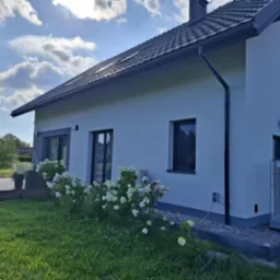 Nowoczesny dom z białą elewacją i szarą podmurówką, widok z ogrodu. Na pierwszym planie kwitnące hortensje i trawnik, w tle błękitne niebo z chmurami.