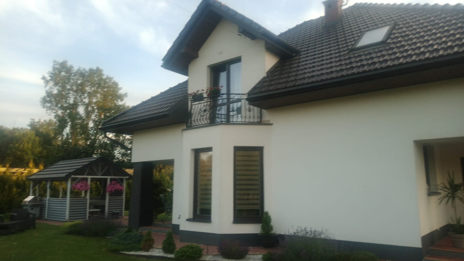 Jasna elewacja domu z balkonem i ciemnymi ramami okiennymi, widoczna altana ogrodowa z kwiatami w tle.