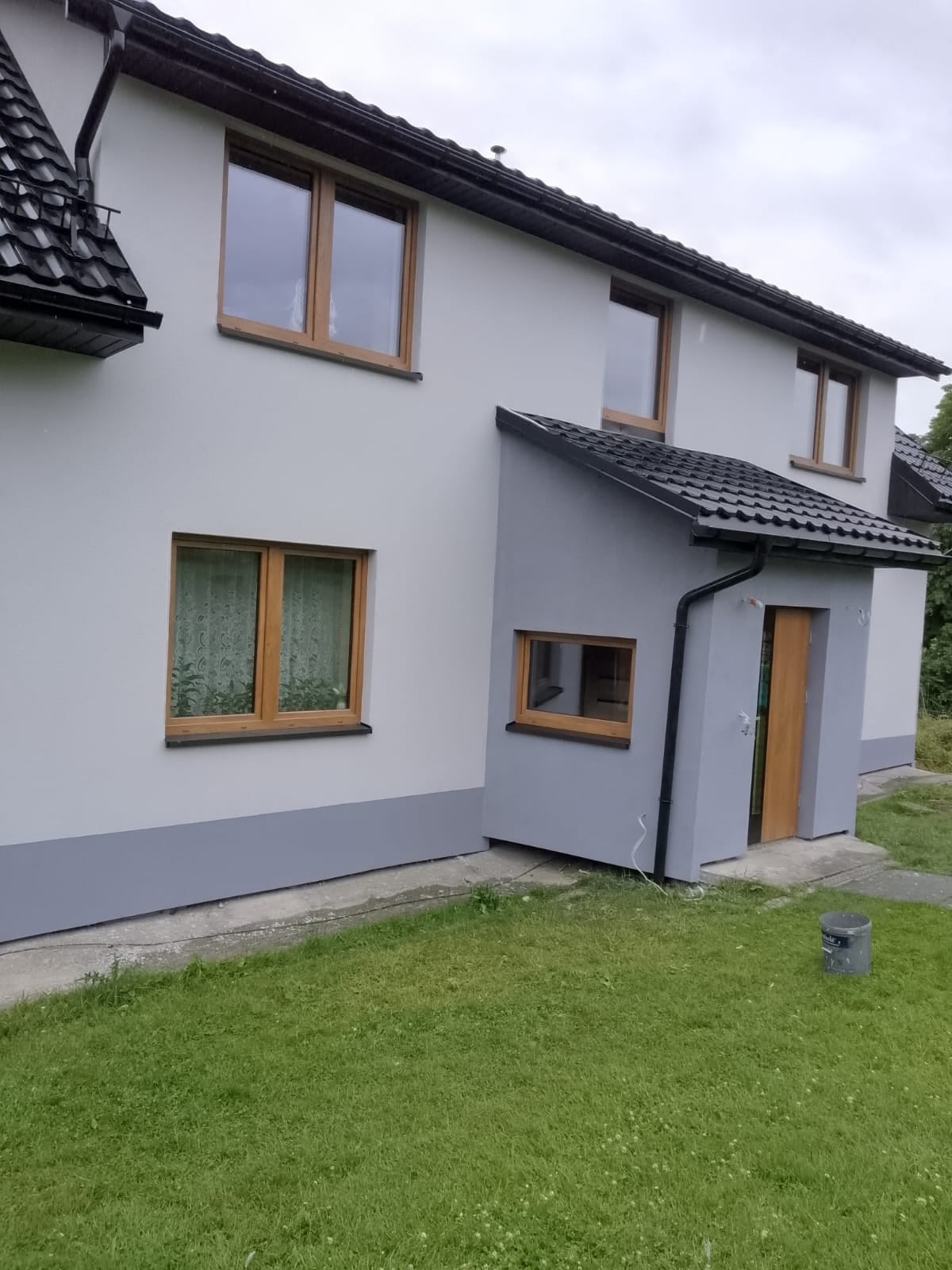 Dwukondygnacyjny dom z jasnoszarą elewacją, brązowymi ramami okiennymi i ciemnoszarą podmurówką, widoczny z poziomu trawnika, z metalowym wiadrem w prawym dolnym rogu.