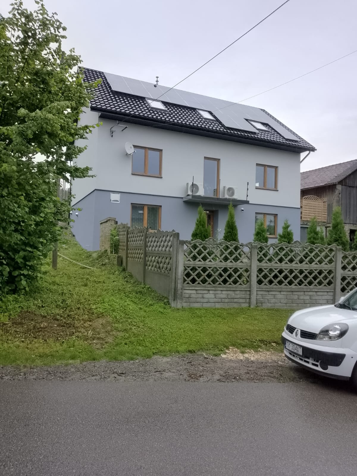 Dwukondygnacyjny dom z szarą elewacją, panelami słonecznymi na dachu i betonowym ogrodzeniem, widok z ulicy w pochmurny dzień.