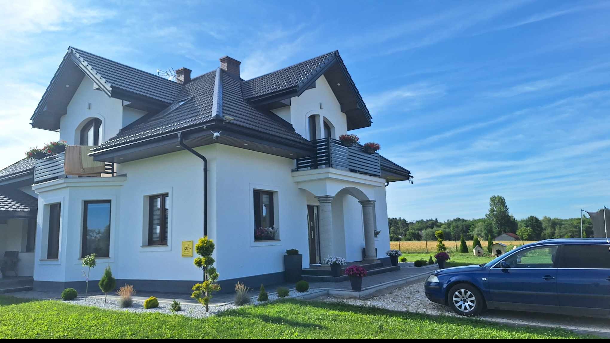 Dwupiętrowy dom z białą elewacją, ciemną dachówką i balkonem z czarną balustradą, widoczny z boku na tle błękitnego nieba i zieleni ogrodu, z zaparkowanym niebieskim kombi.