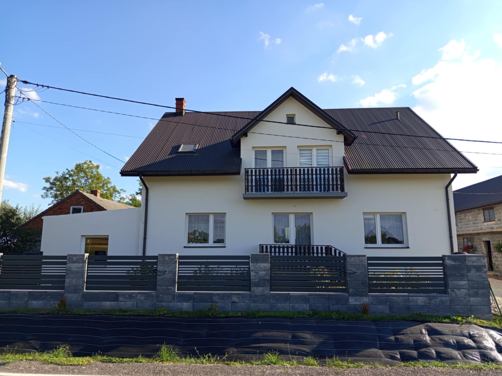 Biały dom z ciemnym dachem i balkonem, ogrodzony szarym płotem z poziomymi panelami. Widok z poziomu ulicy, słoneczny dzień.