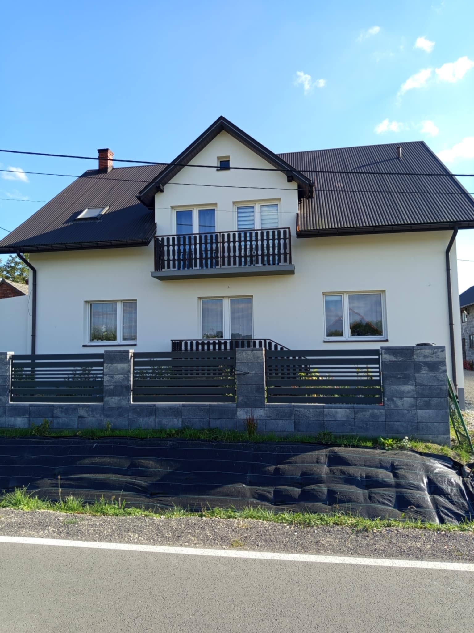 Dwukondygnacyjny dom z białymi ścianami i ciemnym dachem, ogrodzony szarym płotem z betonowych bloczków, na tle błękitnego nieba.