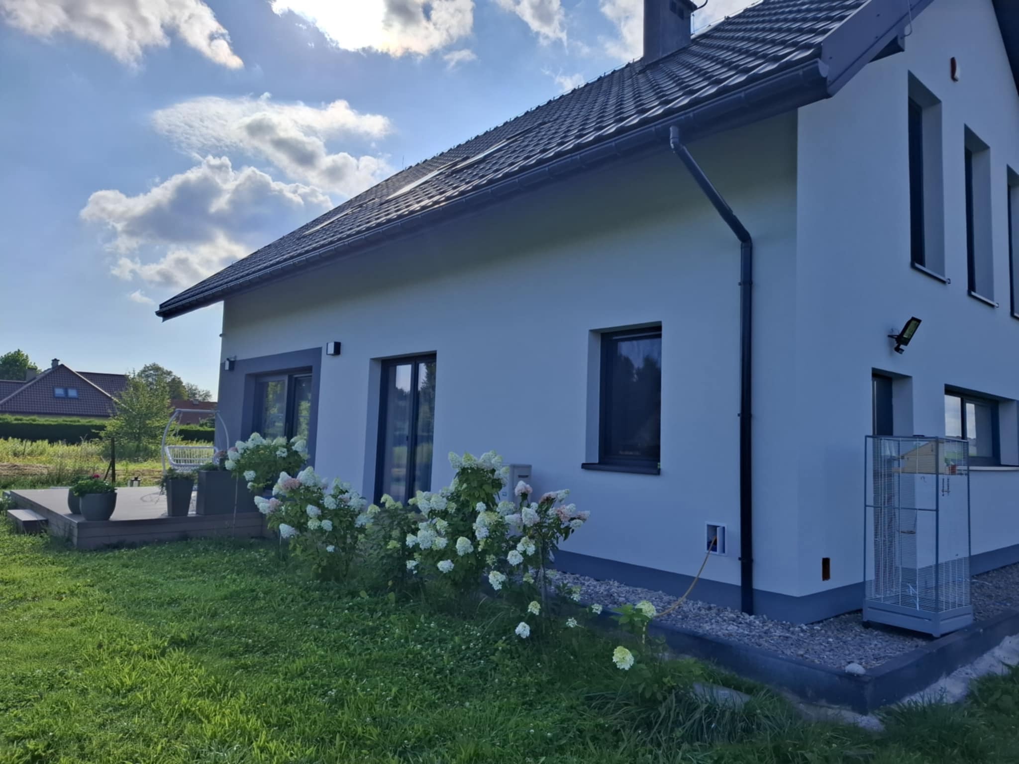 Nowoczesny dom z białą elewacją i szarą podmurówką, widok z ogrodu. Na pierwszym planie kwitnące hortensje i trawnik, w tle błękitne niebo z chmurami.