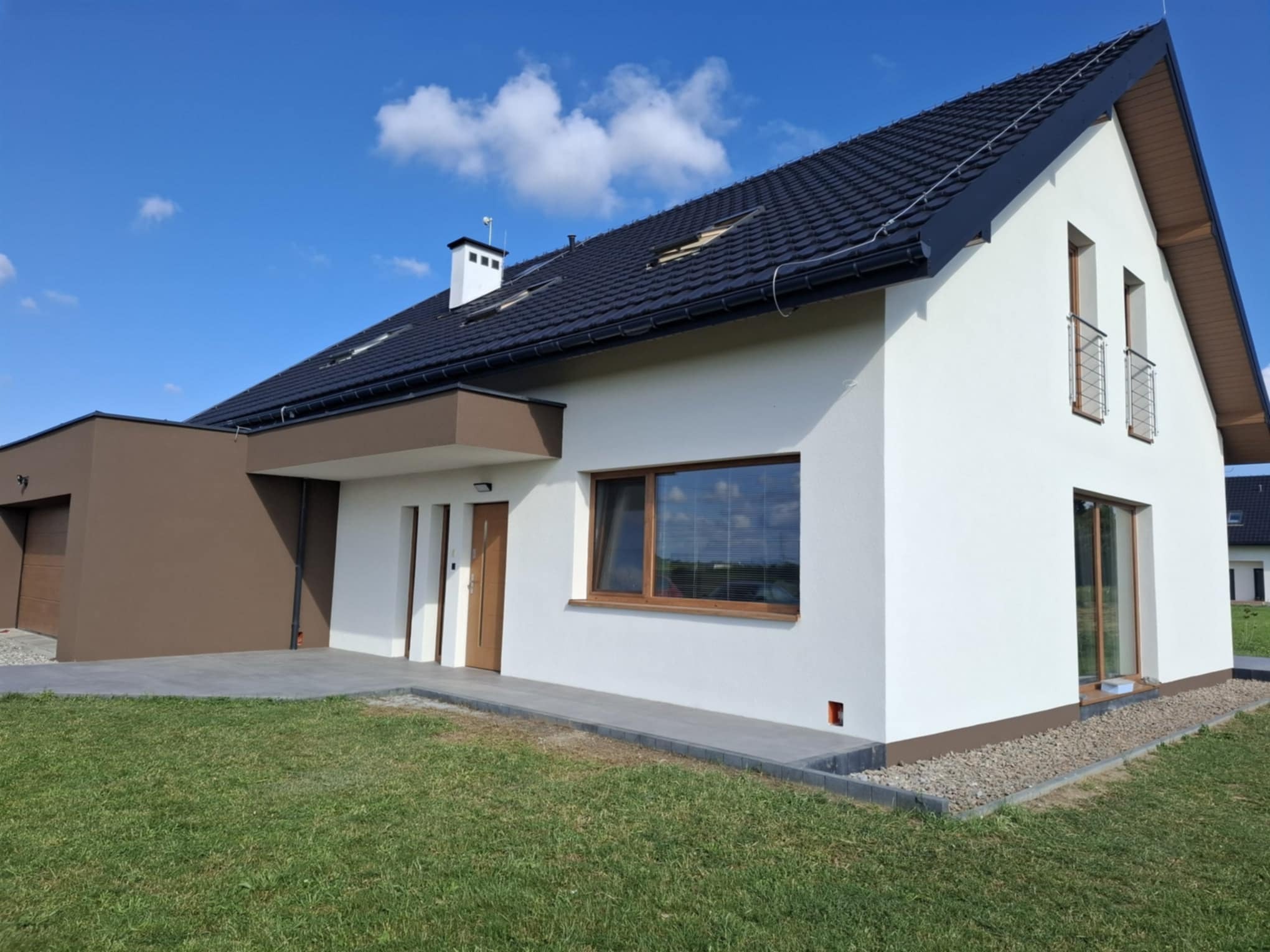 Nowoczesny dom jednorodzinny z białą elewacją i brązowymi akcentami, czarnym dachem i zadbanym trawnikiem w słoneczny dzień. Widok z perspektywy ogrodu.