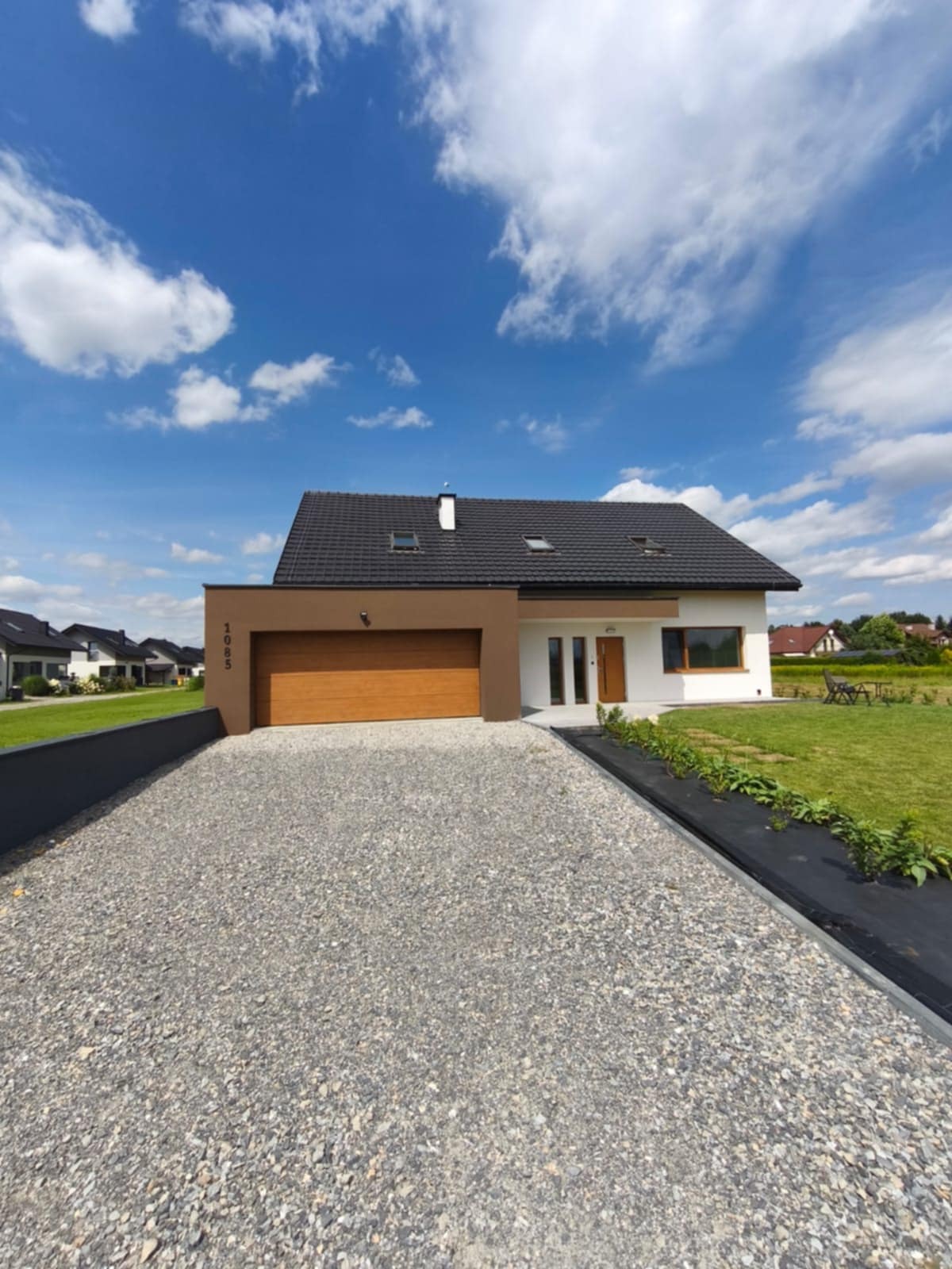 Nowoczesny dom jednorodzinny z brązowym garażem i białymi ścianami, otoczony zielenią i żwirowym podjazdem. Błękitne niebo z chmurami w tle.