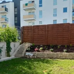 Ogród z drewnianym ogrodzeniem, kamiennym murkiem, schodami i zielonym trawnikiem na tle budynku mieszkalnego.