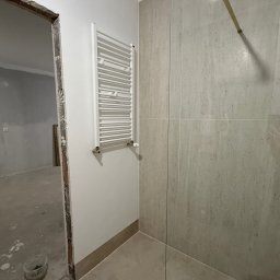 BGS Maciej Bigus - Wykończona łazienka z beżowymi płytkami, grzejnikiem drabinkowym i szklaną kabiną prysznicową. Widoczny surowy otwór drzwiowy do innego pomieszczenia.