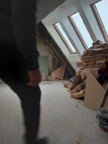 Poddasze w trakcie prac wykończeniowych: widoczne okna dachowe, izolacja z wełny mineralnej, płyty kartonowo-gipsowe i wiadro z zaprawą, zamazana sylwetka osoby w kadrze po lewej.