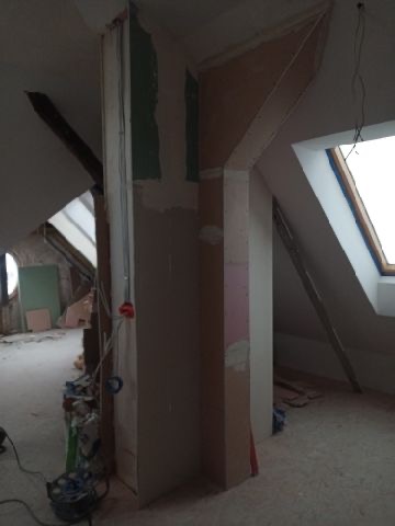 Poddasze w trakcie remontu: widoczne elementy konstrukcyjne dachu, ściany w trakcie szpachlowania i przygotowania do malowania, okno dachowe w ramie.