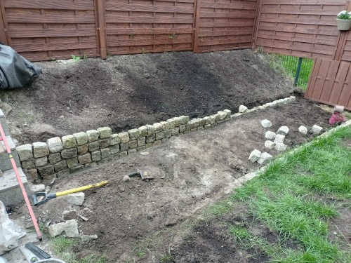Budowa murku z kostki granitowej w ogrodzie, widoczne narzędzia i częściowo ułożona konstrukcja w surowym otoczeniu.