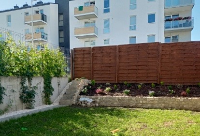 Ogród z drewnianym ogrodzeniem, kamiennym murkiem, schodami i zielonym trawnikiem na tle budynku mieszkalnego.