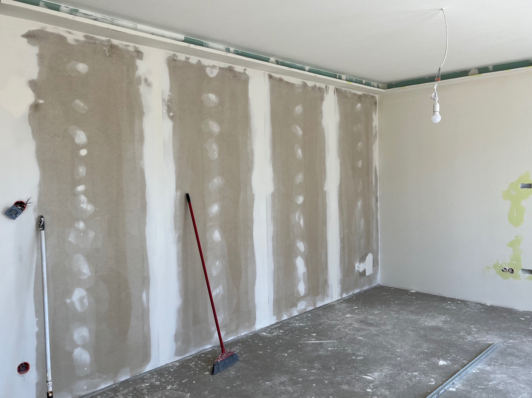 Wnętrze pokoju w trakcie prac wykończeniowych: ściany częściowo pokryte szpachlą, widoczne paski świeżo nałożonej warstwy, na podłodze leży miotła z czerwonym trzonkiem, a z sufitu zwisa żarówka...