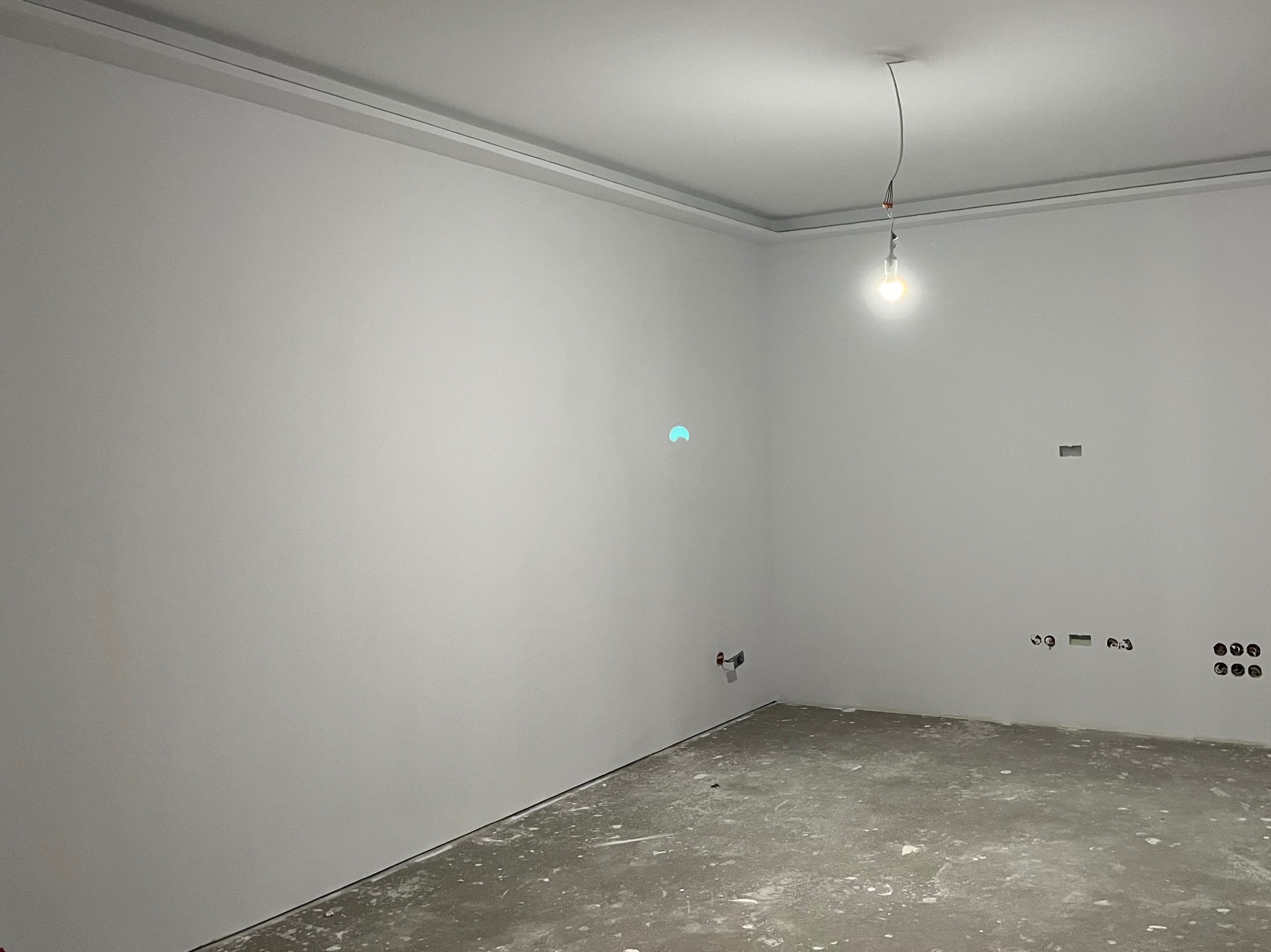 Pusty, jasny pokój w trakcie remontu, świeżo pomalowane ściany na biało, surowa podłoga, widoczne gniazdka elektryczne i wystająca żarówka.