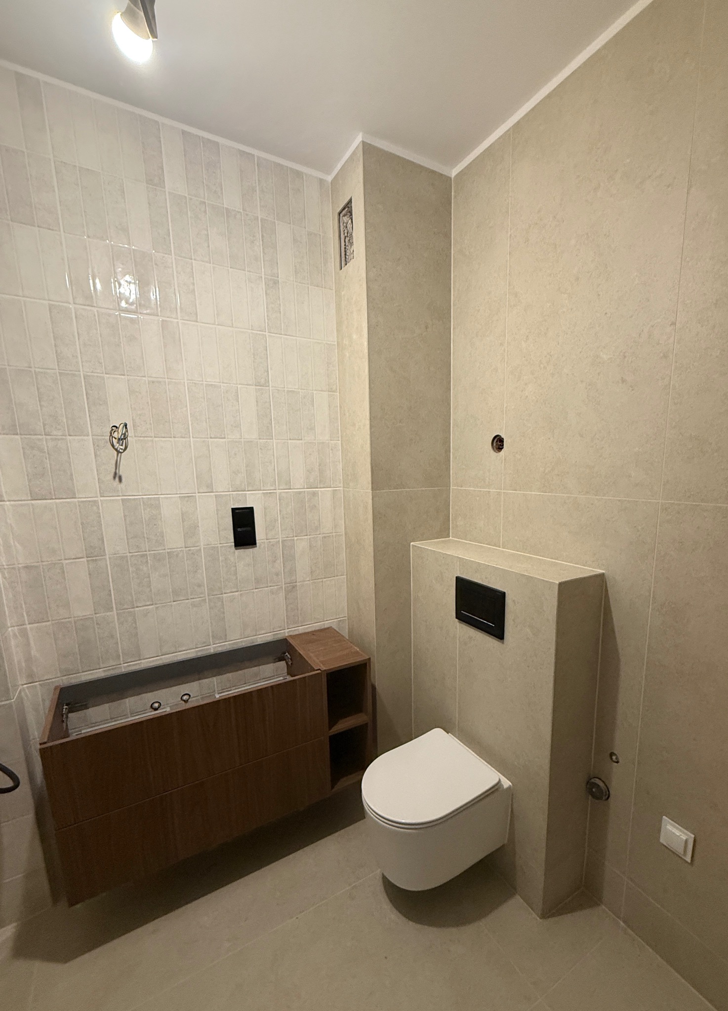 Beżowa łazienka po remoncie: płytki ścienne i podłogowe, podwieszana toaleta, szafka pod umywalkę z drewnianym wykończeniem. Widoczne elementy instalacji.