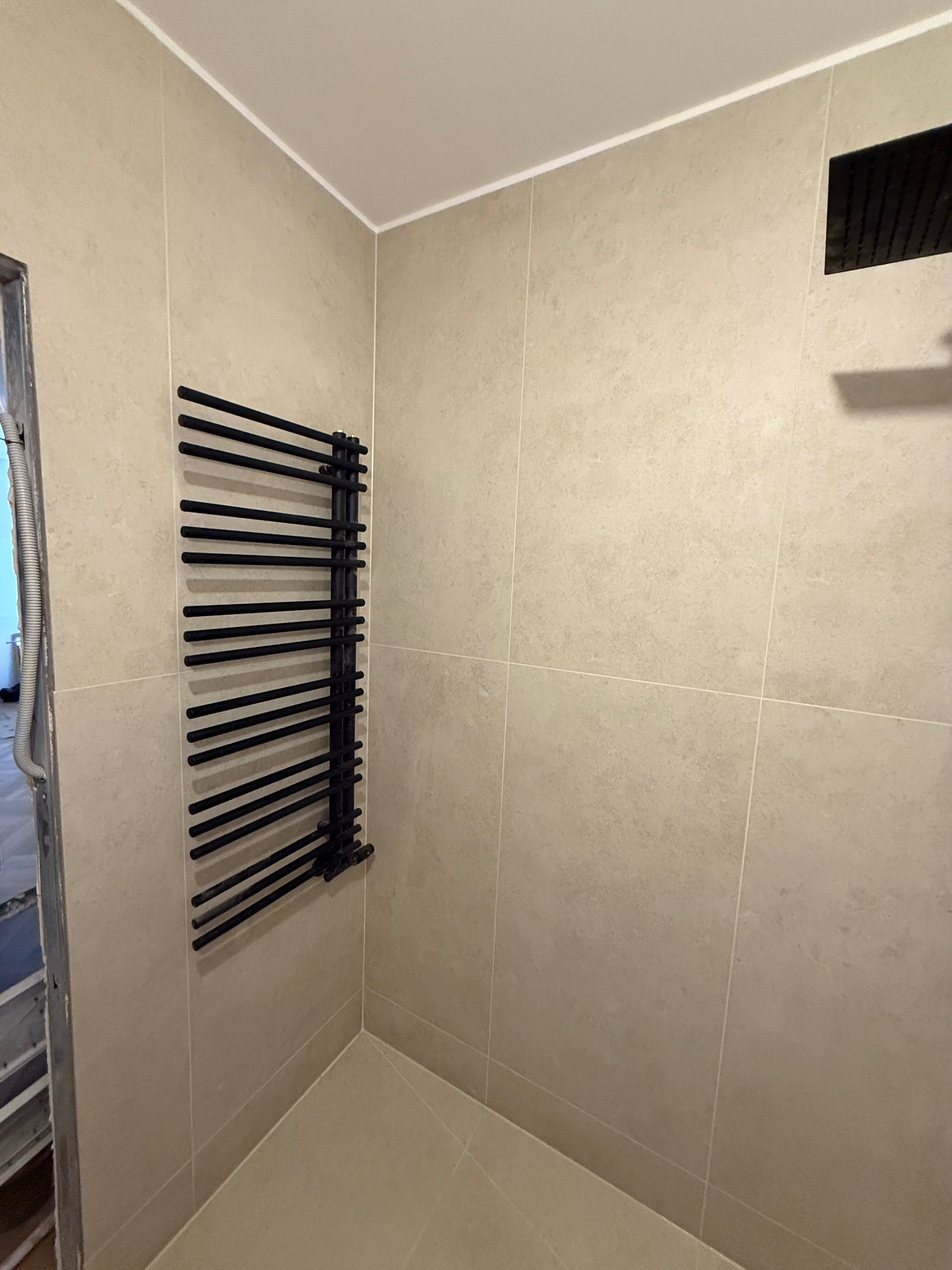 Wykończona łazienka z beżowymi płytkami, czarnym grzejnikiem drabinkowym i deszczownicą. Widoczny fragment futryny drzwi.