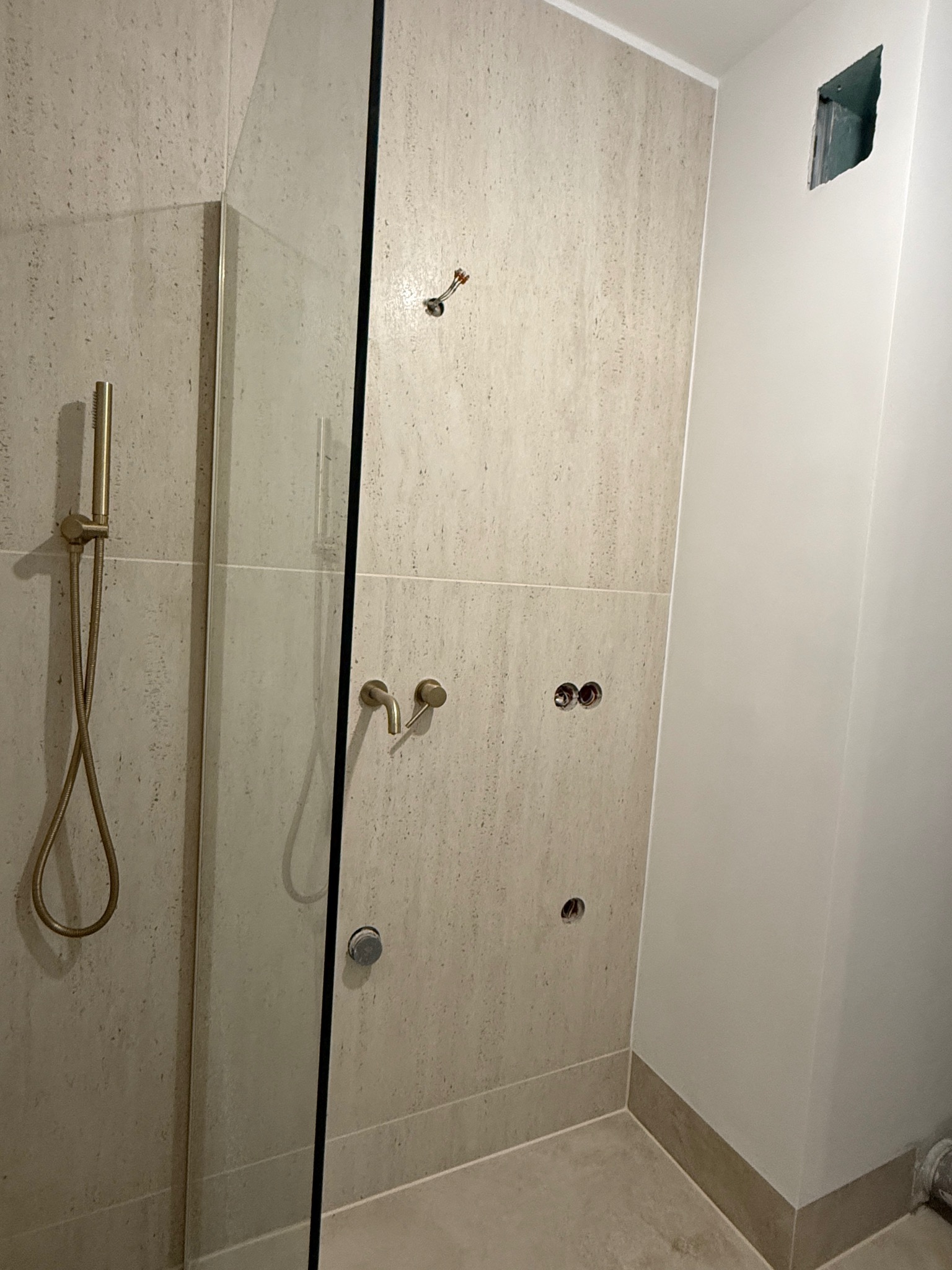 Niedokończona łazienka z beżowymi płytkami imitującymi kamień, widoczne przyłącza do baterii, otwór w ścianie i szklana kabina prysznicowa z złotym zestawem natryskowym.