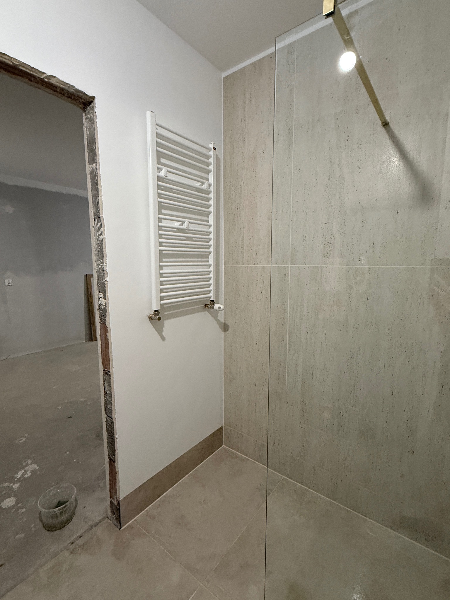 Wykończona łazienka z beżowymi płytkami, grzejnikiem drabinkowym i szklaną kabiną prysznicową. Widoczny surowy otwór drzwiowy do innego pomieszczenia.