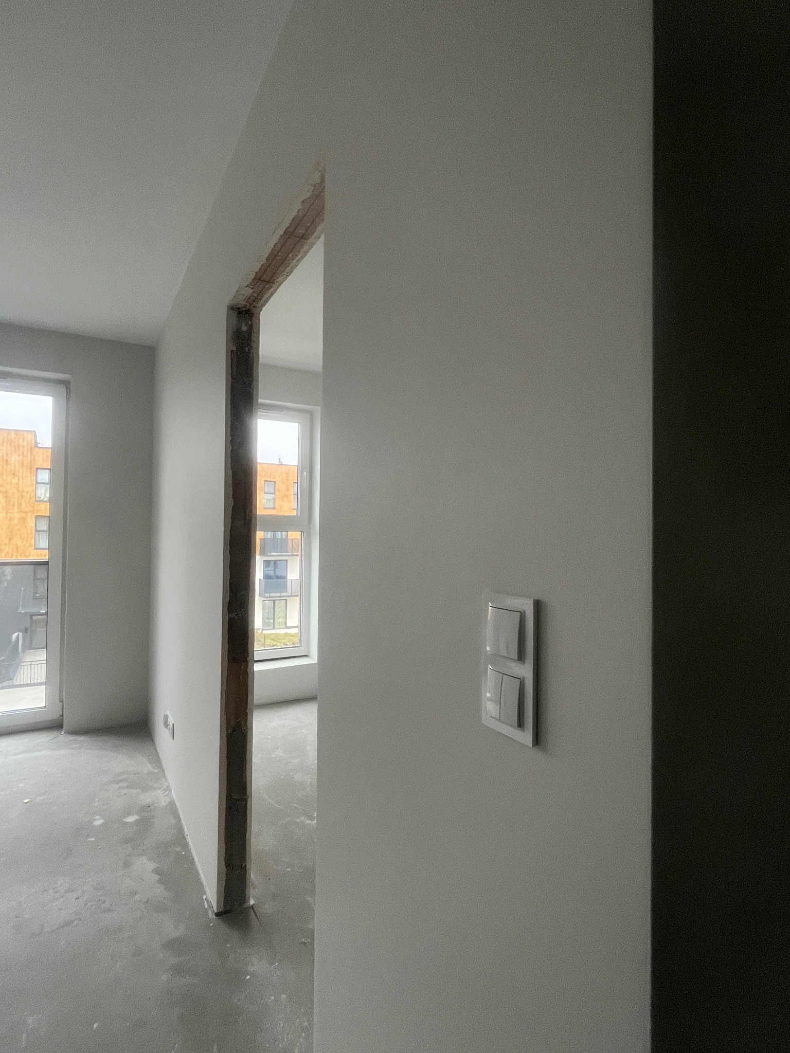 Wnętrze w trakcie remontu: gładkie, pomalowane ściany w odcieniach szarości, surowy beton na podłodze, widoczne okna i fragmenty konstrukcji ściany działowej.