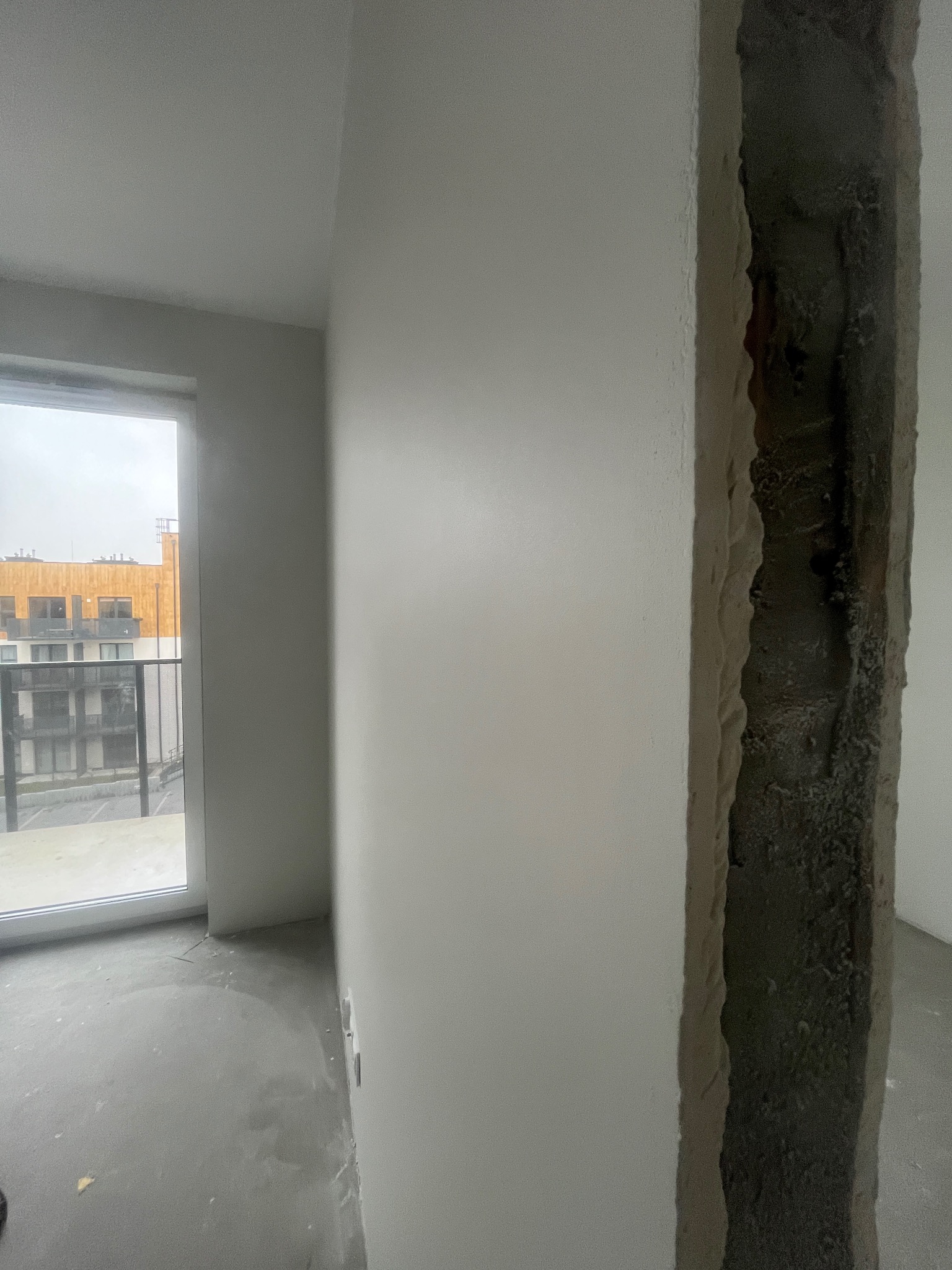Wnętrze w trakcie remontu: gładka, pomalowana ściana sąsiaduje z surowym, betonowym filarem. Widoczne okno z widokiem na budynek. Szary beton na podłodze.